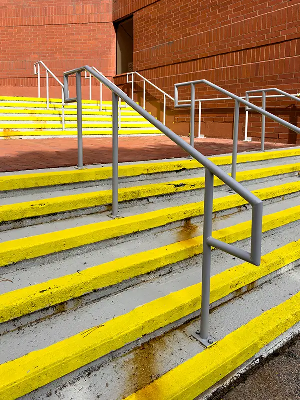 The outside entrance to a large red brick building with multiple concrete steps. The wide stairs have yellow paint on the edge of each step. There are galvanized metal handrails in the middle.