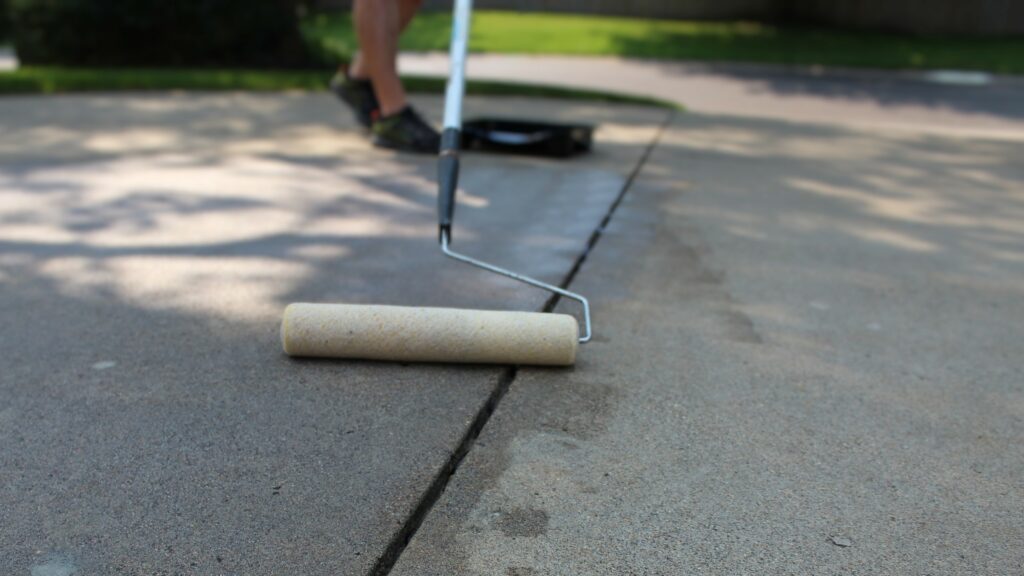 A person is using a roller brush to paint sealer on a concrete slab driveway